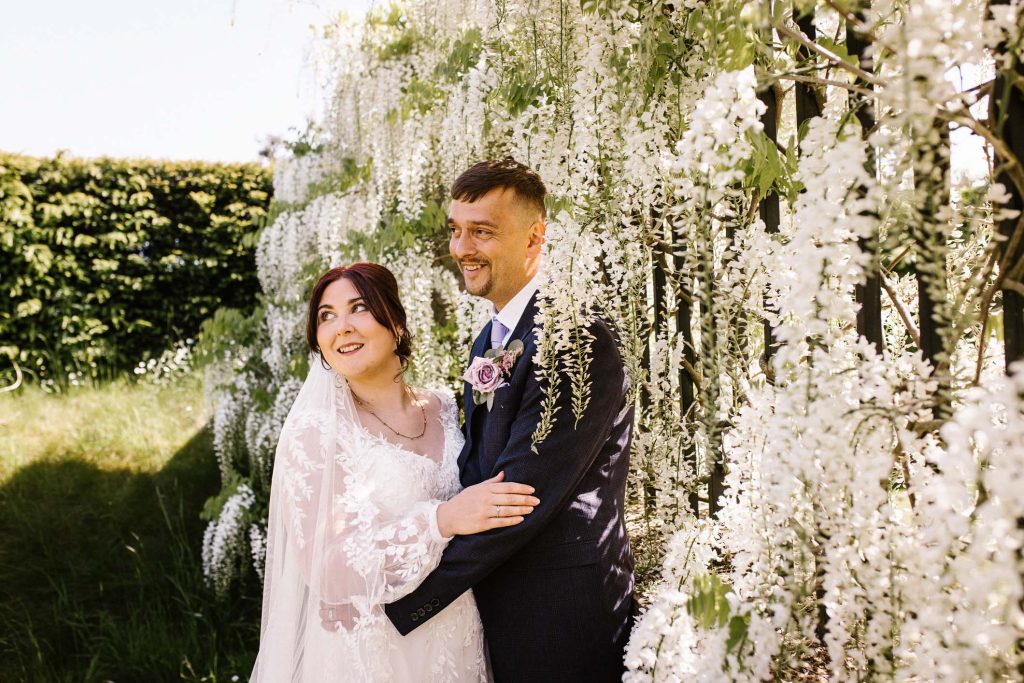 Bride and groom holding each other in a close embrace e, looking away, smiling, surrounded by a wall of white wisteria flowers. Spring time at an English manor house garden.