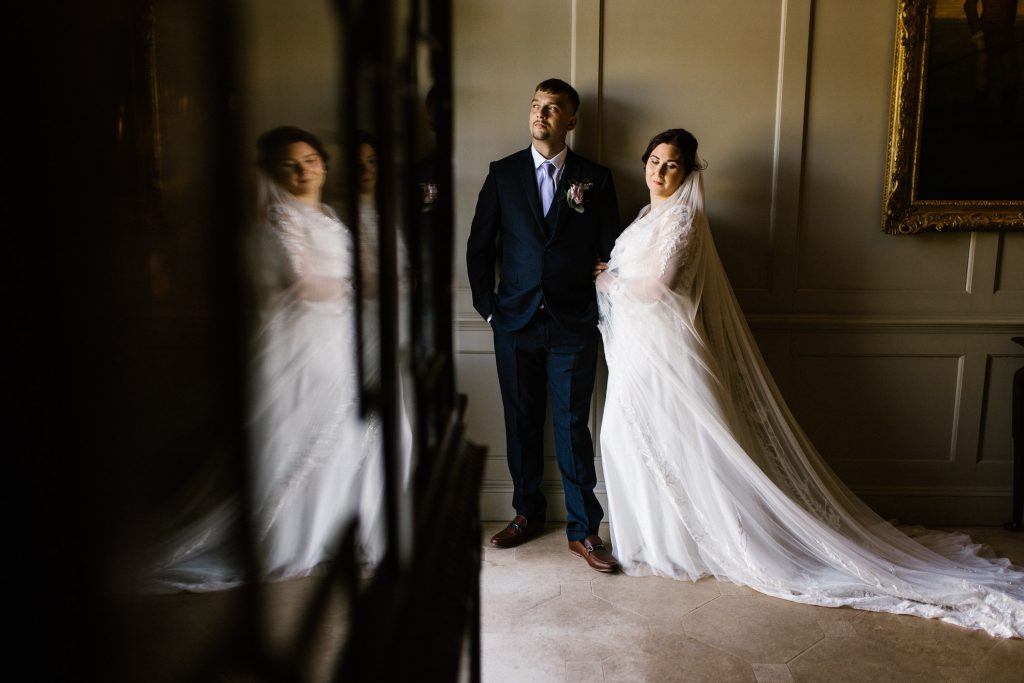 Bride and groom standing inside an English Manor House, there is a reflection of the bride in a glass front cabinet on the left.