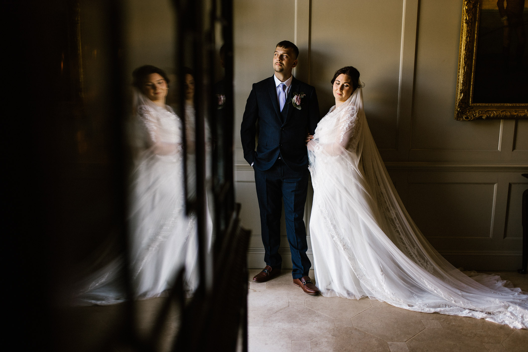 Wedding couple standing together in a Manor House corridor. Elegant interior with cream walls and hanging lights. Bride and groom holding hands