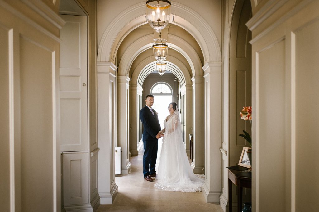 Wedding couple standing together in a Manor House corridor. Elegant interior with cream walls and hanging lights. Bride and groom holding hands