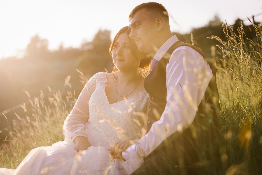 Golden hour light falling on the valley, bride and groom sitting on the ground, with grooms arm around his bride. Looking into distance with peaceful expressions. Spring time, sunset vibe